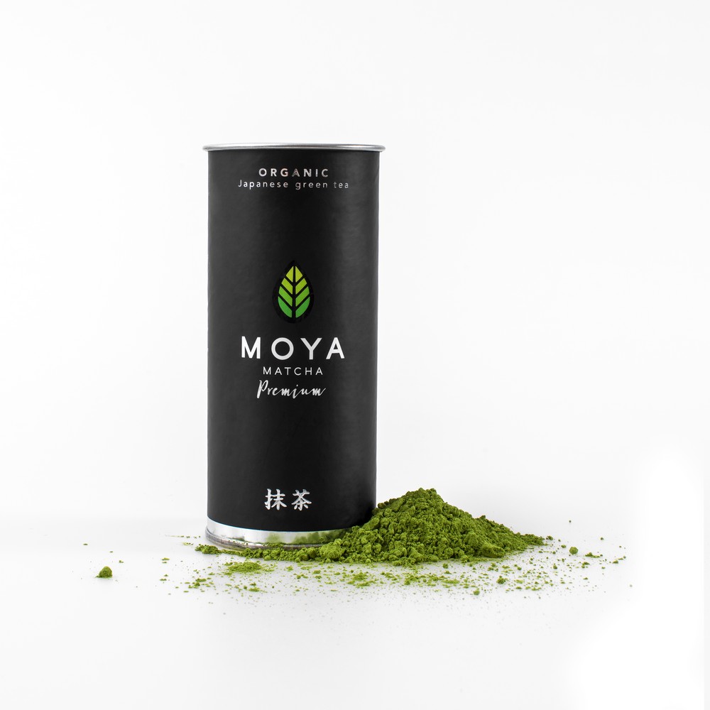 Moya Organikus Prémium Matcha Zöld Tea fekete hengeres dobozban, elöl zöld matchaporral fehér háttéren.