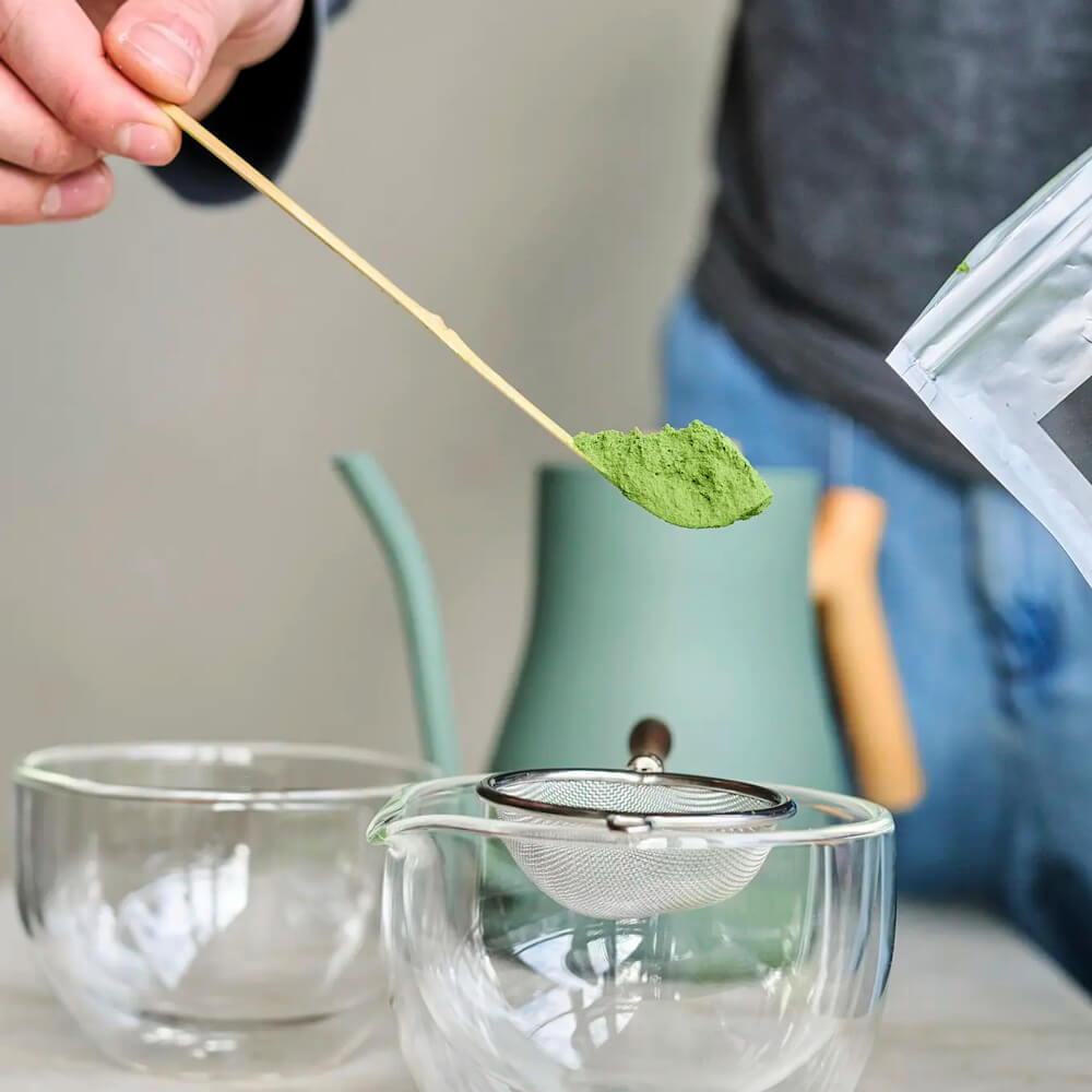 Világos bambusz matcha teakanál használat közben, zöld matchaporral üvegtál és szita felett.