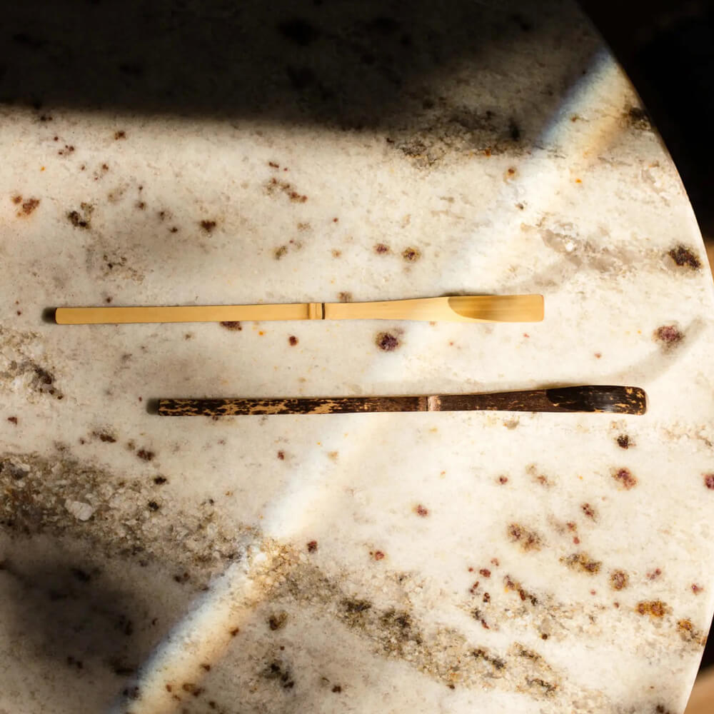 Világos bambusz matcha teakanál és egy sötét teakanál felülnézetben, márvány mintás felületen.