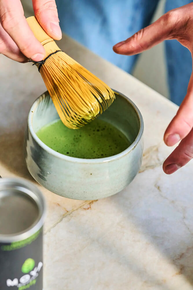 Matcha tea és bambusz habverő szett használat közben, élénk zöld ital kerámia tálban, közelről fotózva