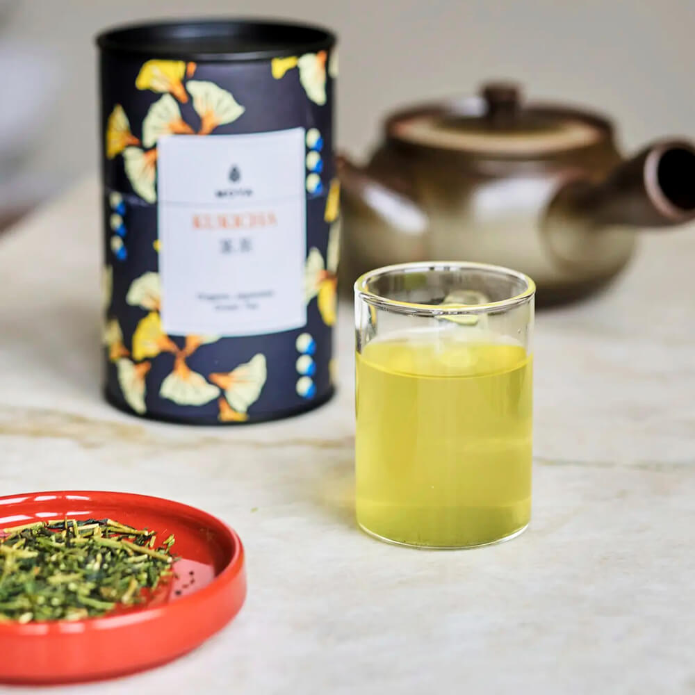 Moya Kukicha organikus japán zöld tea doboz, üvegpohár tea és tealevelek piros tálkában