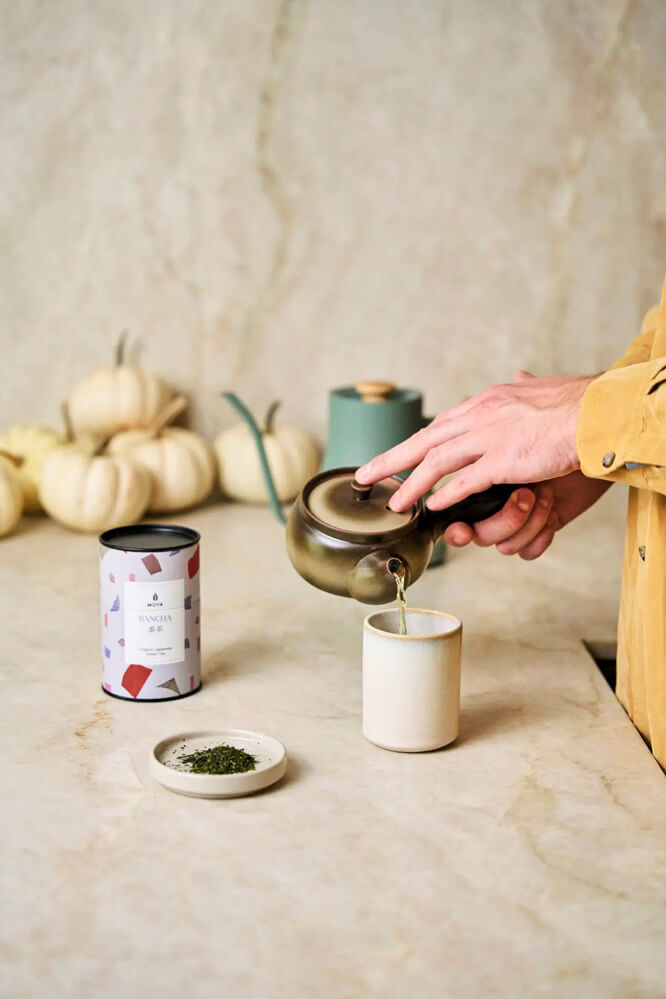 Moya Bancha 60 g organikus japán zöld tea lifestyle fotó, teáskannából bögrébe töltve, a dobozzal a pulton.