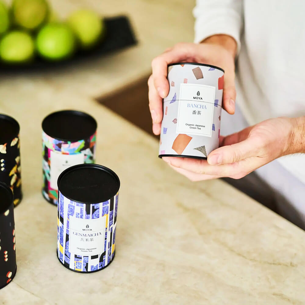 Moya Bancha 60 g organikus japán zöld tea díszdoboz kézben, konyhapulton, más MOYA teásdobozok mellett.