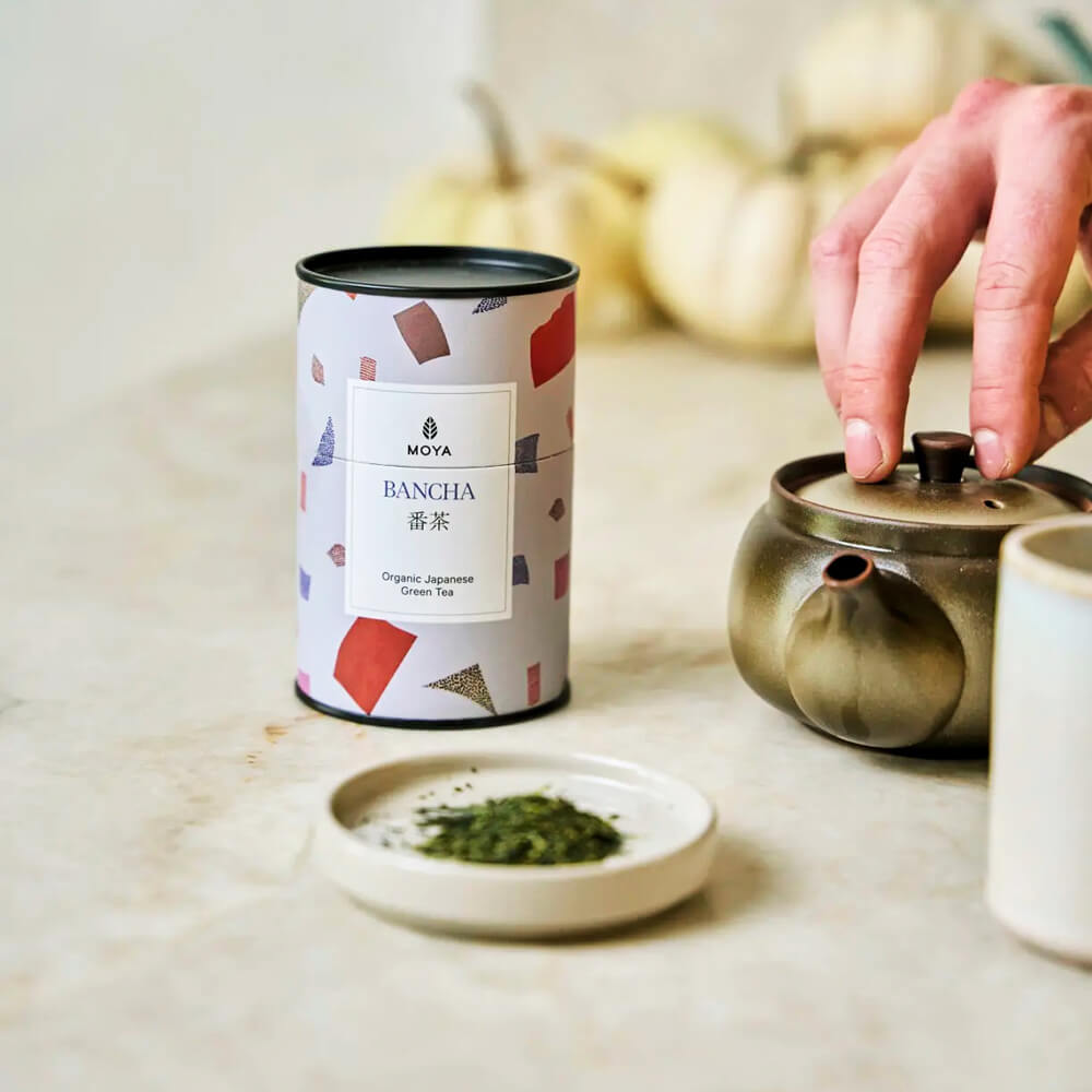 Moya Bancha 60 g organikus japán zöld tea doboza teáskannával és szálas teával, konyhapulton elrendezve.