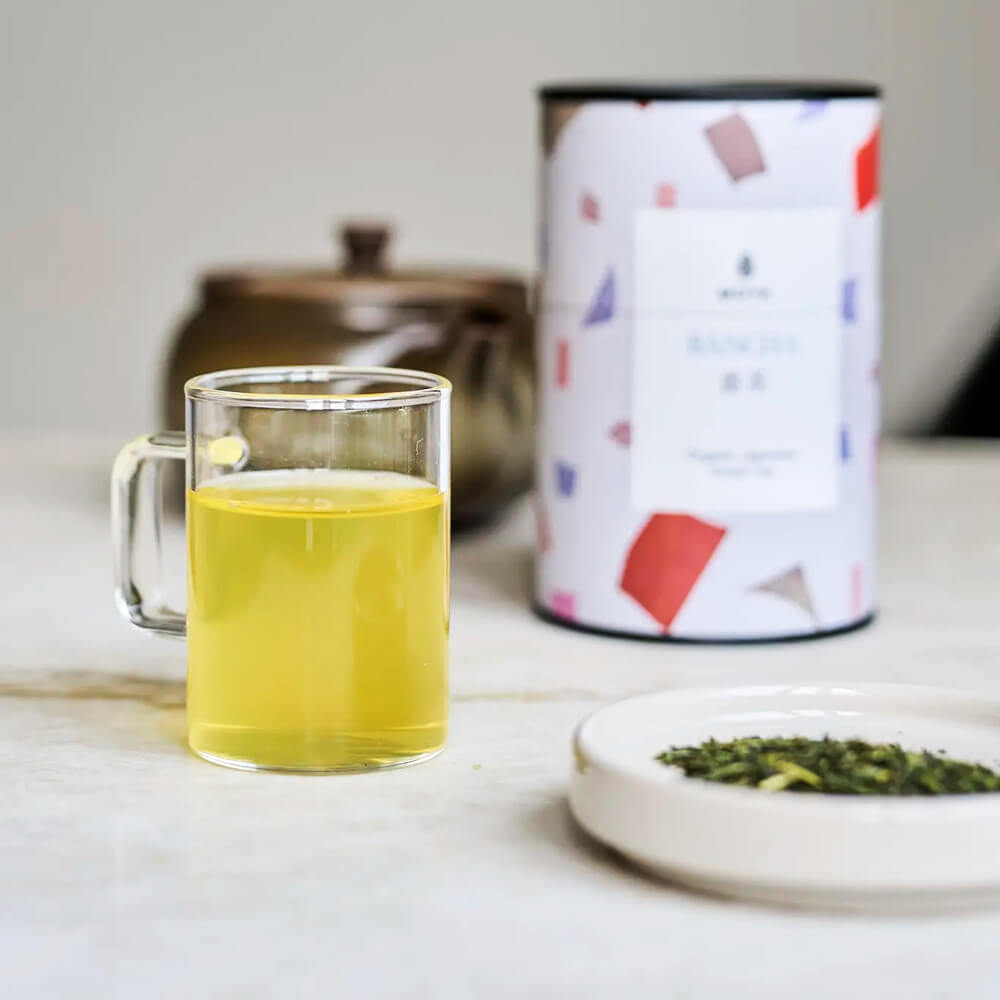 Moya Bancha 60 g organikus japán zöld tea főzve üvegbögrében, mellette szálas tea és a doboz a háttérben.