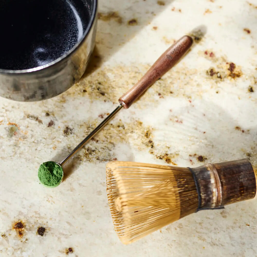 Matcha Adagolókanál matcha porral márvány lapon, mellette bambusz habverő és tál részlete