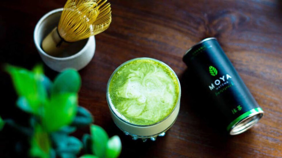 Matcha latte felülnézetből zöld örvénnyel, mellette Moya doboz az asztalon