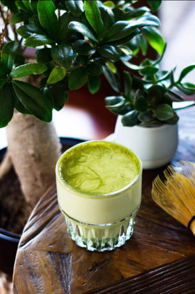 Matcha latte üvegpohárban faasztalon, mellette chasen és növény