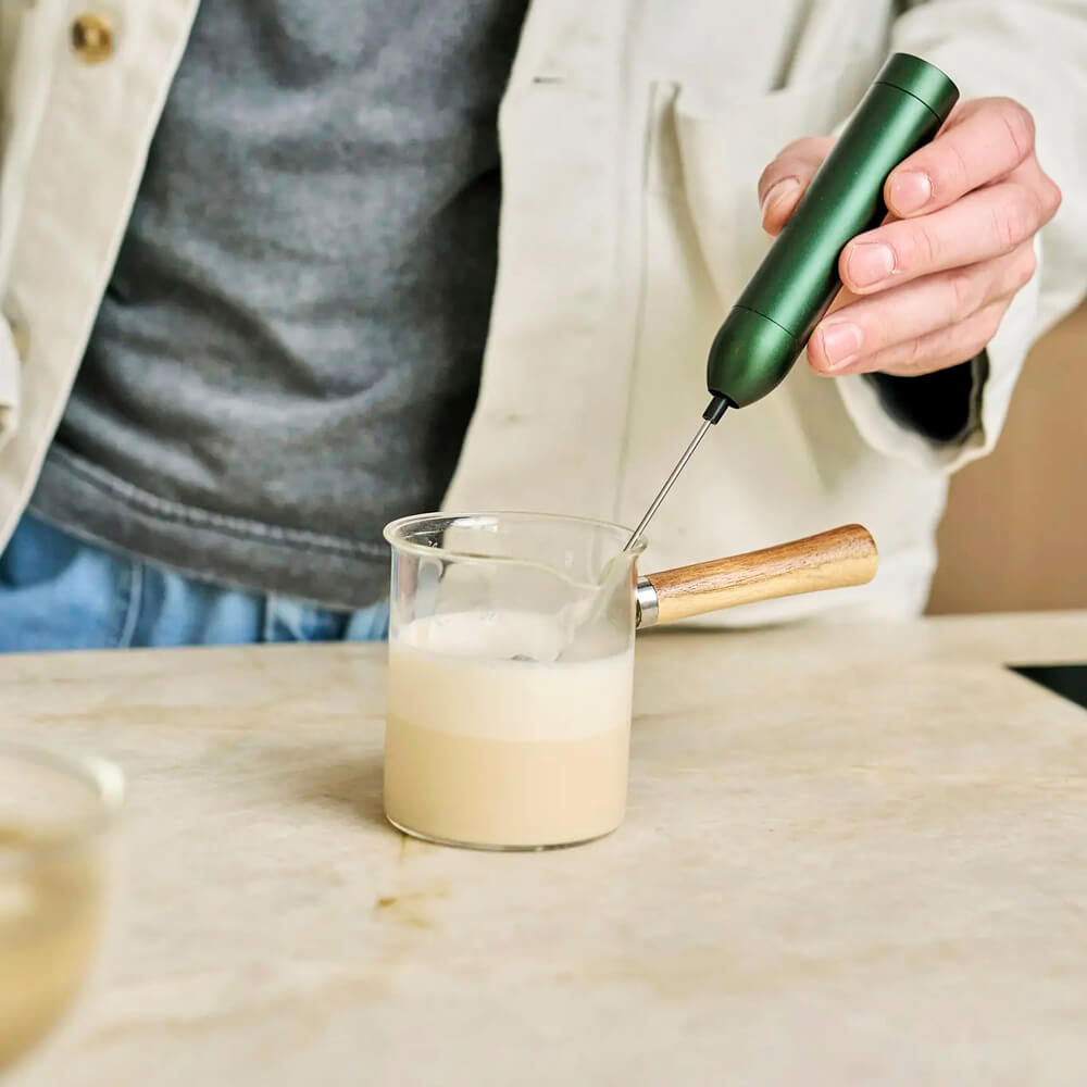 Moya Organikus Hojicha Por ital készítése tejjel: kézi habosító pohárban, üveg kiöntőben a konyhapulton