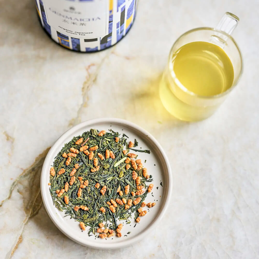 Moya Genmaicha organikus japán zöld tea levelei tálkában, üvegpohár teával és dobozzal felülnézetből