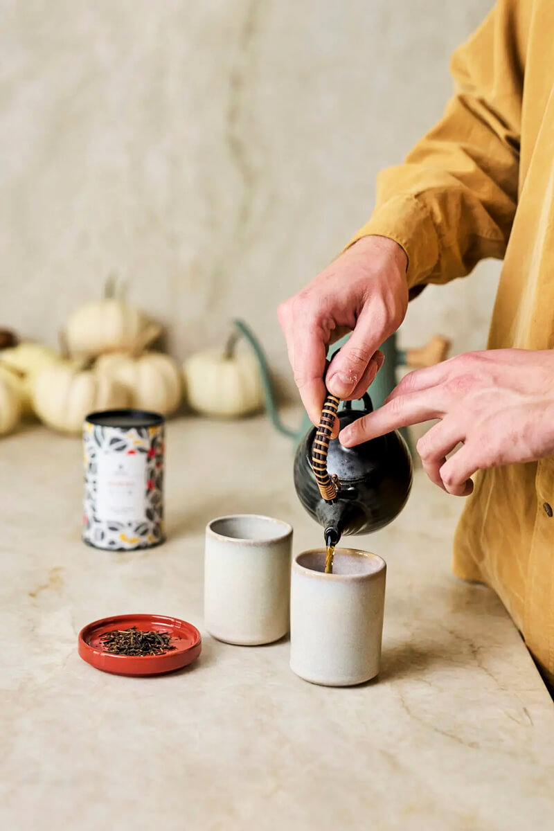 Moya Hojicha organikus japán zöld tea kiöntés közben, fekete teáskannával, két csészével és tealevelekkel.