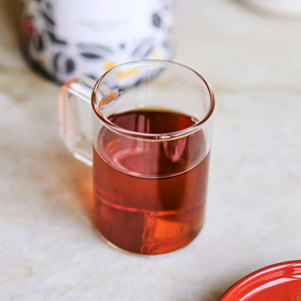 Moya Hojicha organikus japán zöld tea egy üveg bögrében, borostyánszínű itallal és tealevelekkel.