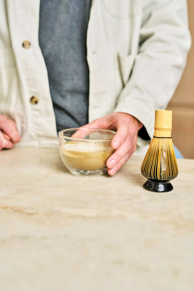 Krémes hojicha latte üvegpohárban, mellette chasen tartó az asztalon