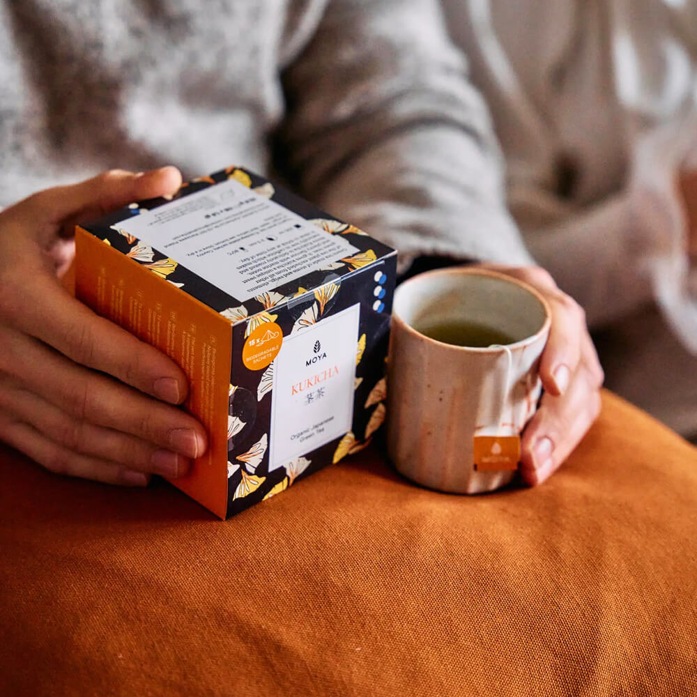 Moya Kukicha organikus japán filteres zöld tea doboz és bögre kézben, otthonos lifestyle fotón