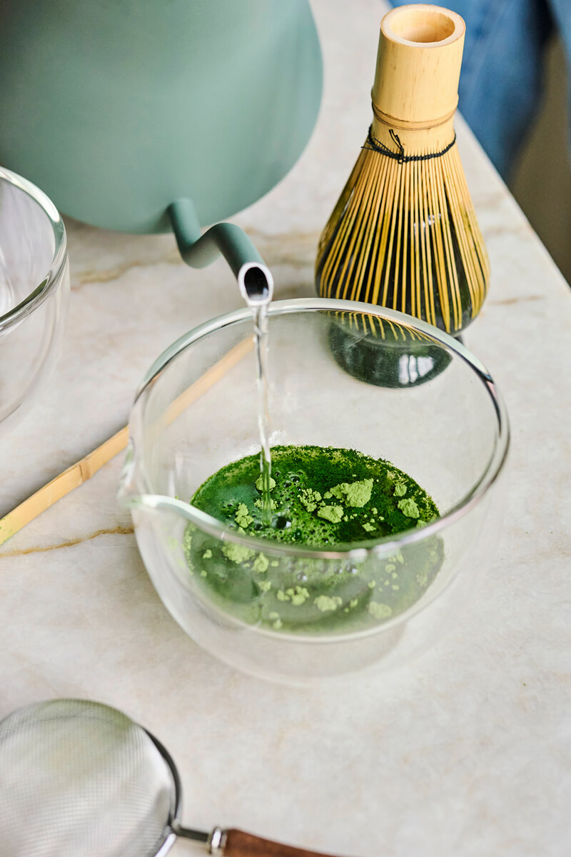 Víz öntése matcha porhoz egy kerámia tálban bambusz habverő mellett