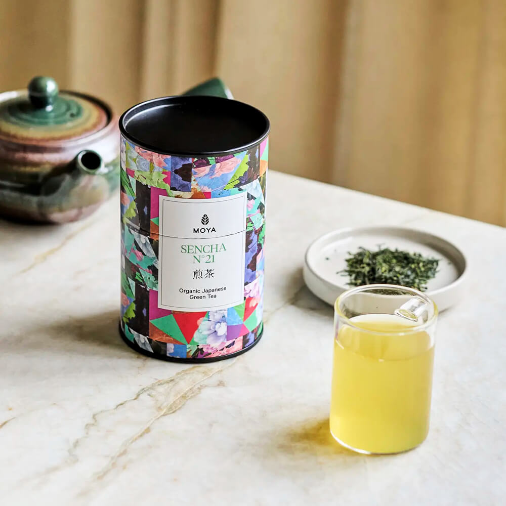 Moya Sencha No. 21 organikus japán zöld tea doboz, üvegpohár tea és tealevelek enteriőr fotón