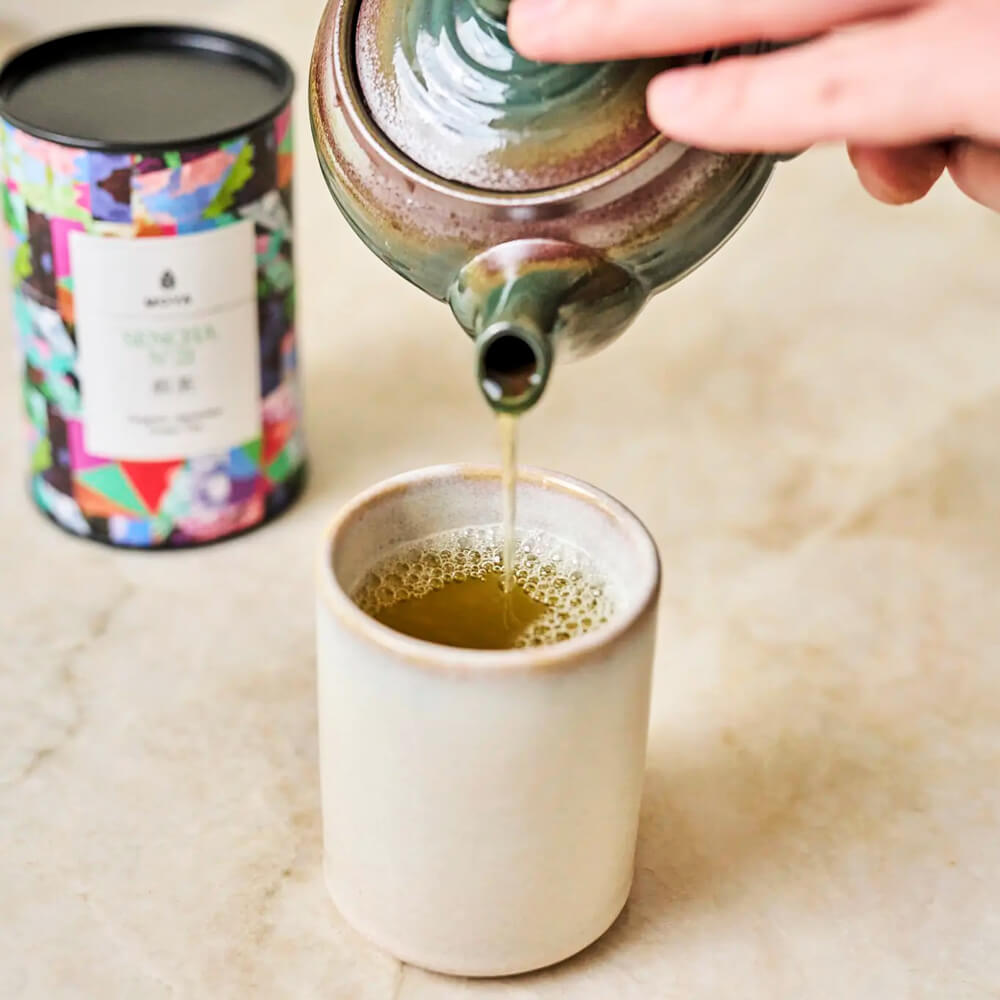 Moya Sencha No. 21 organikus japán zöld tea kitöltése csészébe teáskannából, a doboz a háttérben látszik