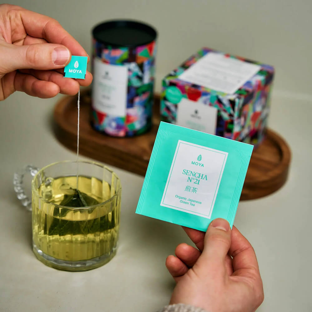 Moya Sencha No. 21 organikus japán zöld tea filtertasak kézben, üvegbögrével és dobozzal lifestyle fotón