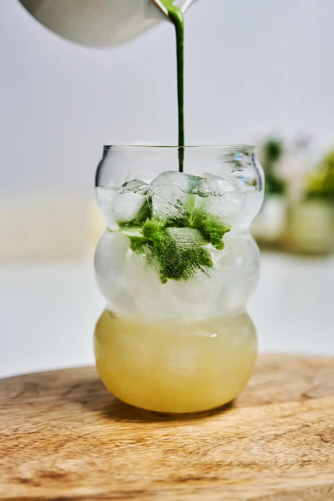 Matcha-Yuzu-Tonic-recept-friss,-citrusos-és-gyors Matcha Yuzu Tonic recepthez matchát öntenek jéggel teli pohárba, alul világos yuzu-tonic réteggel.