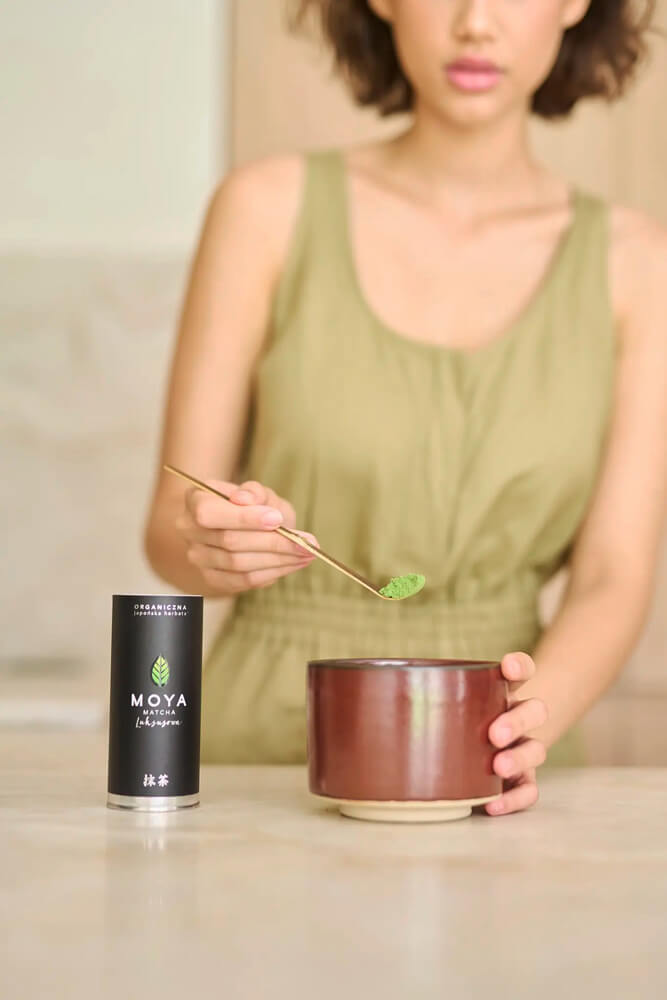 Matcha por adagolása barna csészébe, mellette Moya Matcha csomagolás