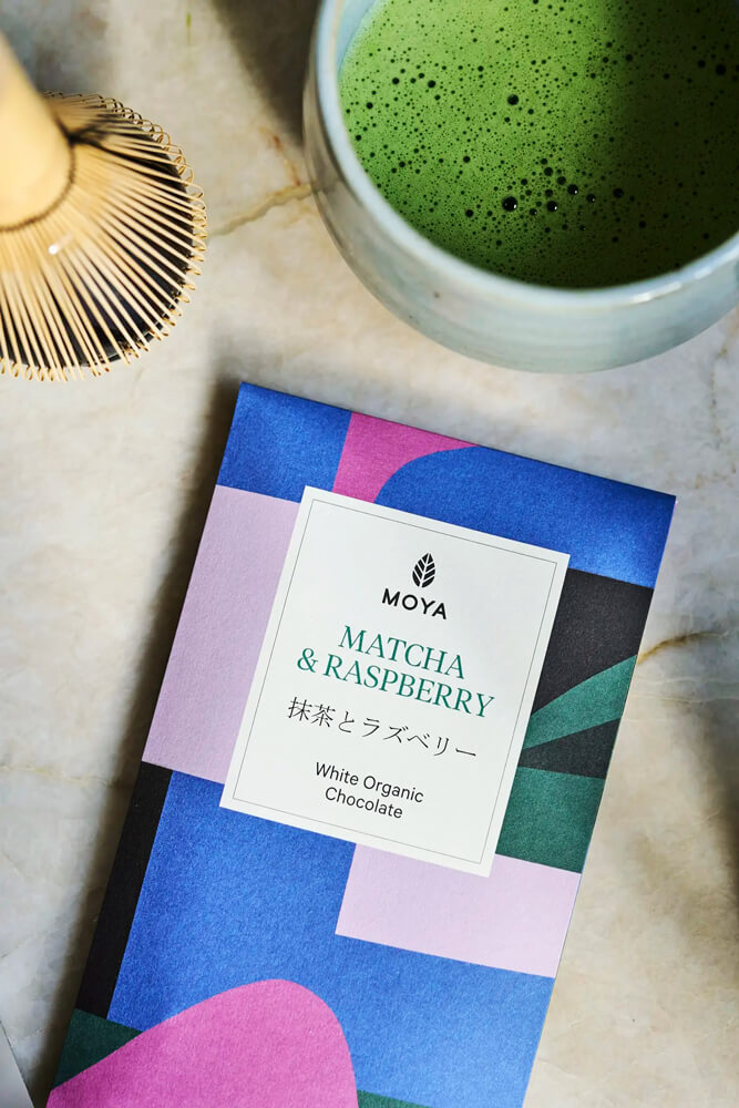 Moya Matcha Fehércsokoládé Málnával – csomagolás Moya Matcha Fehércsokoládé Málnával 80 g csomagolása matcha teával és bambusz ecsettel felülnézetből.