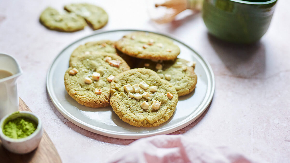 Matcha süti fehér csokoládéval tányéron, matcha porral és zöld bögrével lifestyle fotón.