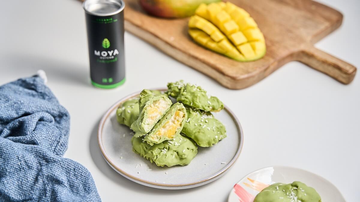 Mangófalatok matcha csokoládéban tálalva Matcha csokoládéba mártott mangófalatok tányéron, mellette Moya matcha doboz és friss mangó