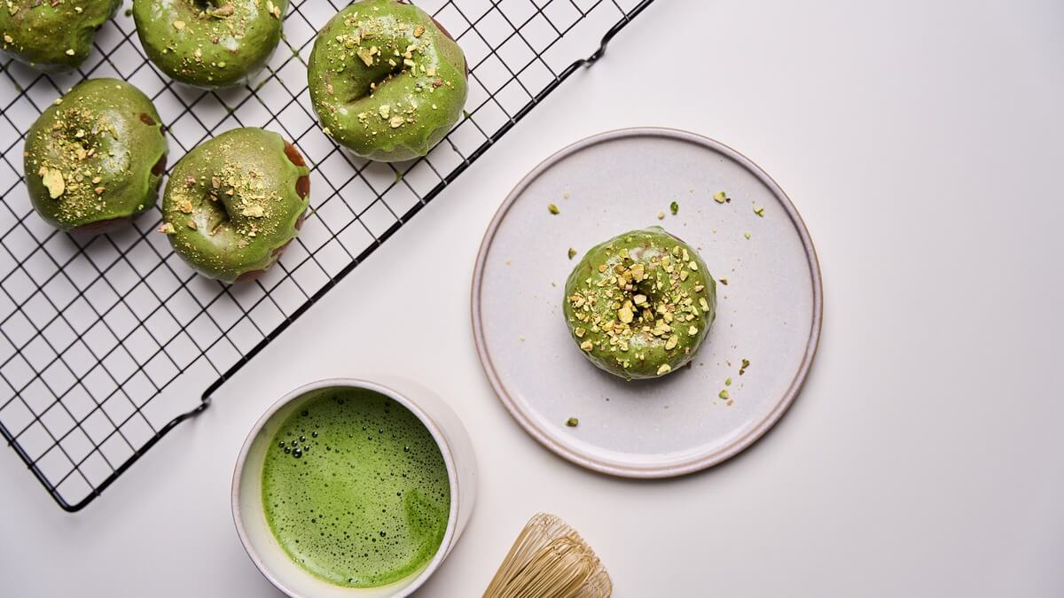 Matcha fánkok mázzal Matchás mázzal bevont kelt fánkok rácson és tányéron, mellette felvert matcha chasennel