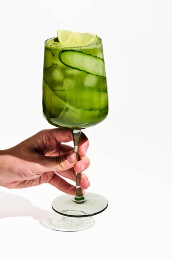 Kézben tartott talpas pohár alkoholmentes matcha koktéllal, uborkaszalaggal és lime-mal